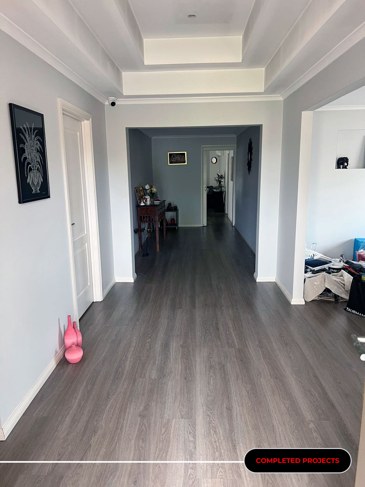 Long, light-gray hallway with layered ceiling panels, grey wood floor, and a pink vase on the left.